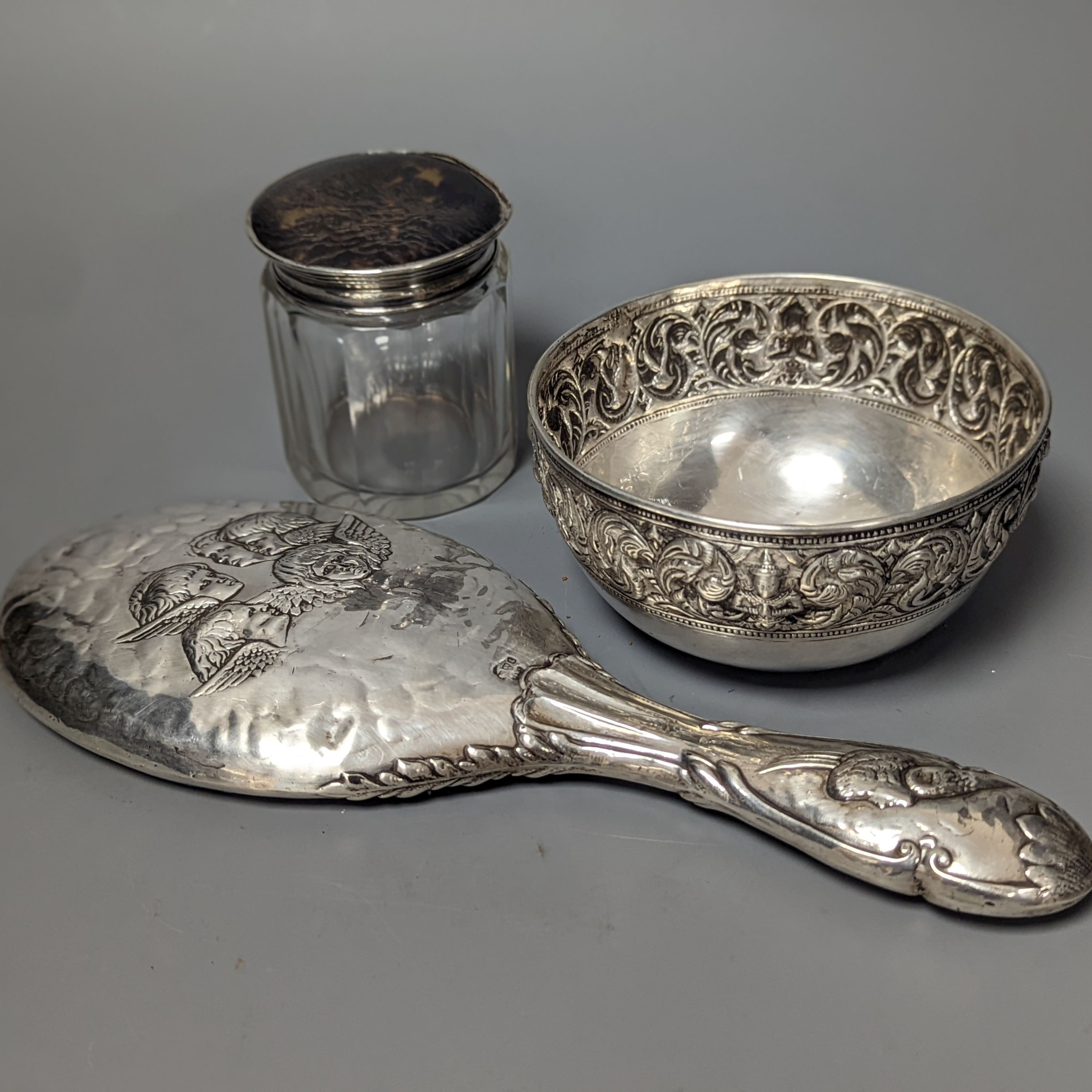 An Edwardian silver mounted 'Reynold's Angels' hand mirror, a silver and tortoiseshell lidded glass toilet jar and a Thai? white metal bowl.
